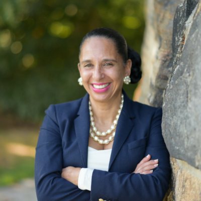 Adriana Leela Bohm wearing a navy suit with a pearl necklace and matching earrings, with her arms crossed and hair pulled back, smiling