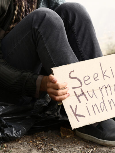 Person sitting on ground with cardboard sign that says "Seeking Human Kindness."