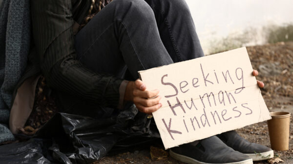 Person sitting on ground with cardboard sign that says "Seeking Human Kindness."
