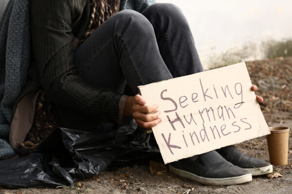 Person sitting on ground with cardboard sign that says "Seeking Human Kindness."