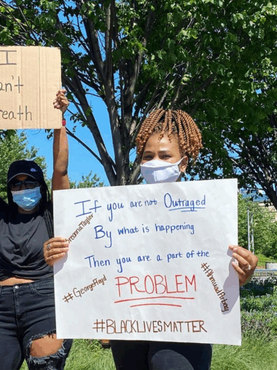Two women holding signs at police violence protest, standing in front of Wilmington Police Department SUV on Martin Luther King Blvd in Wilmington