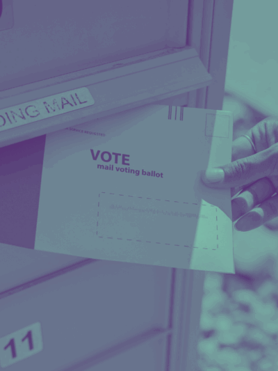 photograph of a person's hand putting a mail ballot in an "outgoing mail" mailbox.