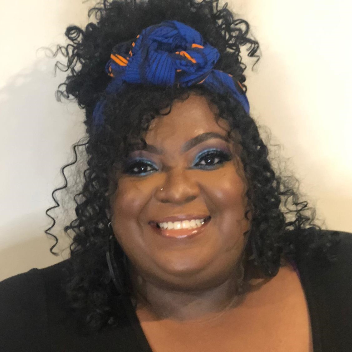 A headshot of Nyemade Boiwu wearing her curly hair half-up, half-down, a black shirt, and a royal blue scarf in her hair with orange details.