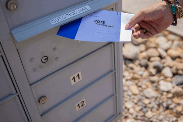 Person putting a vote by mail ballot into an outgoing mail post office box outside in daylight.