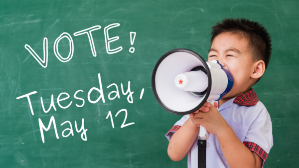 A kid with a megaphone stands in front of a chalkboard that reads "Vote! Tuesday, May 12"