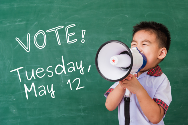 A kid with a megaphone stands in front of a chalkboard that reads "Vote! Tuesday, May 12"