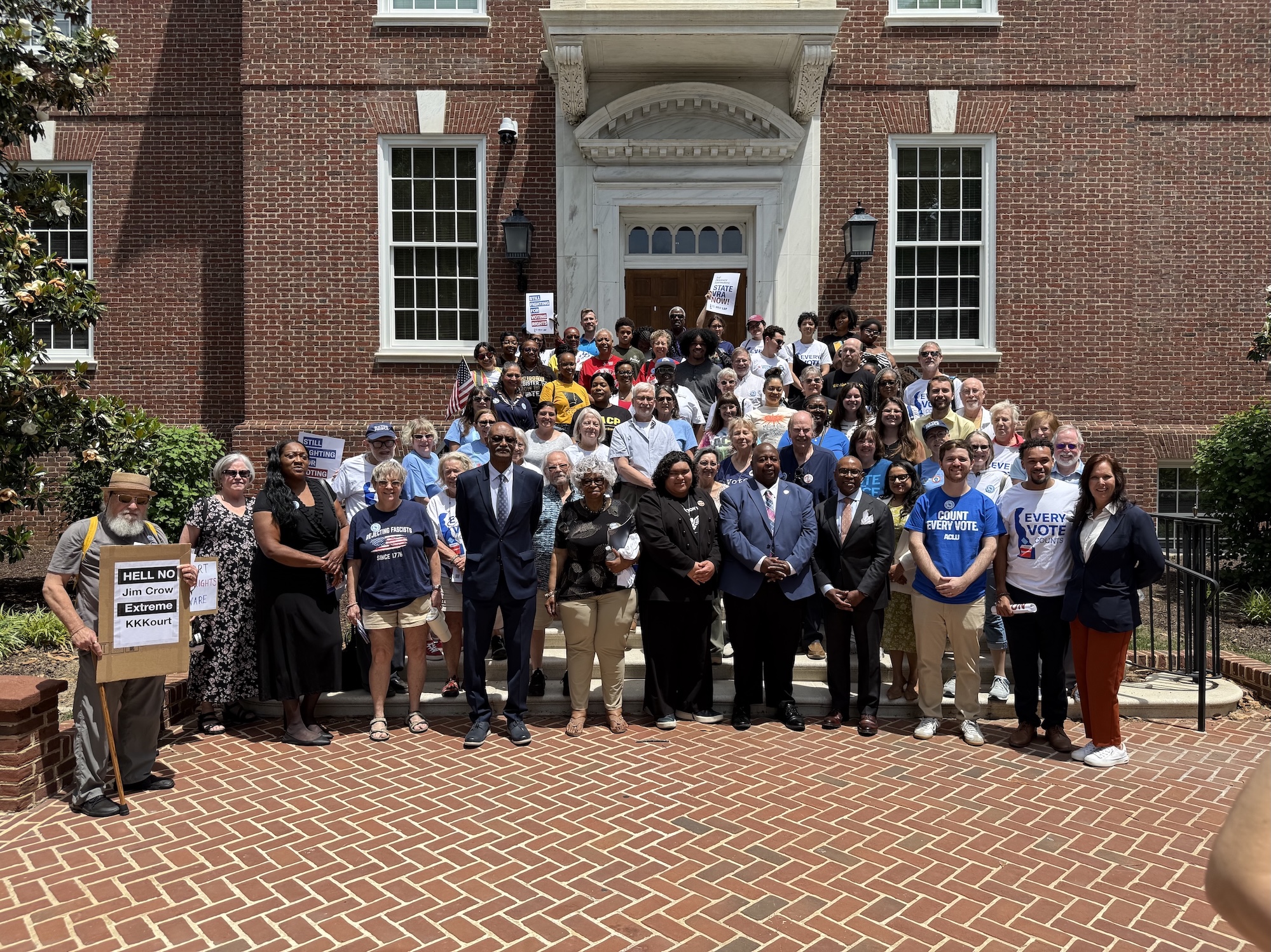 photograph of a large group of people standing on the steps of Legislative Hall in Dover, some holding signs, with the speakers of the event in the front