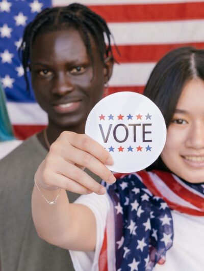 four young people standing in front of an American flag, one person holding up a sticker that says "VOTE"