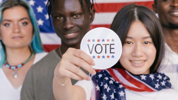 four young people standing in front of an American flag, one person holding up a sticker that says "VOTE"