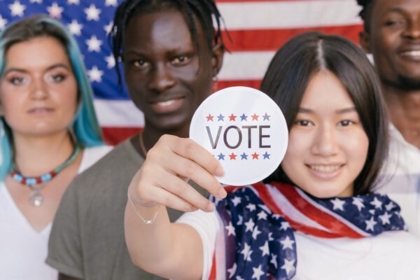 four young people standing in front of an American flag, one person holding up a sticker that says "VOTE"