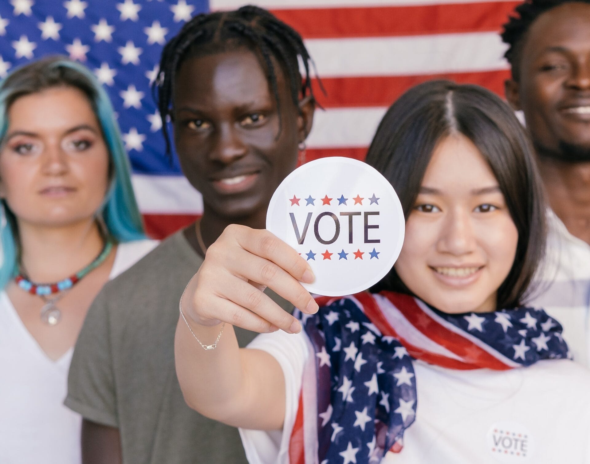 four young people standing in front of an American flag, one person holding up a sticker that says "VOTE"
