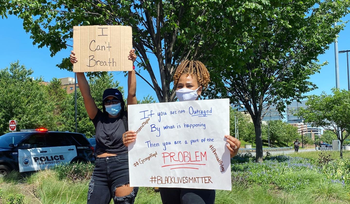 Two women holding signs at police violence protest, standing in front of Wilmington Police Department SUV on Martin Luther King Blvd in Wilmington