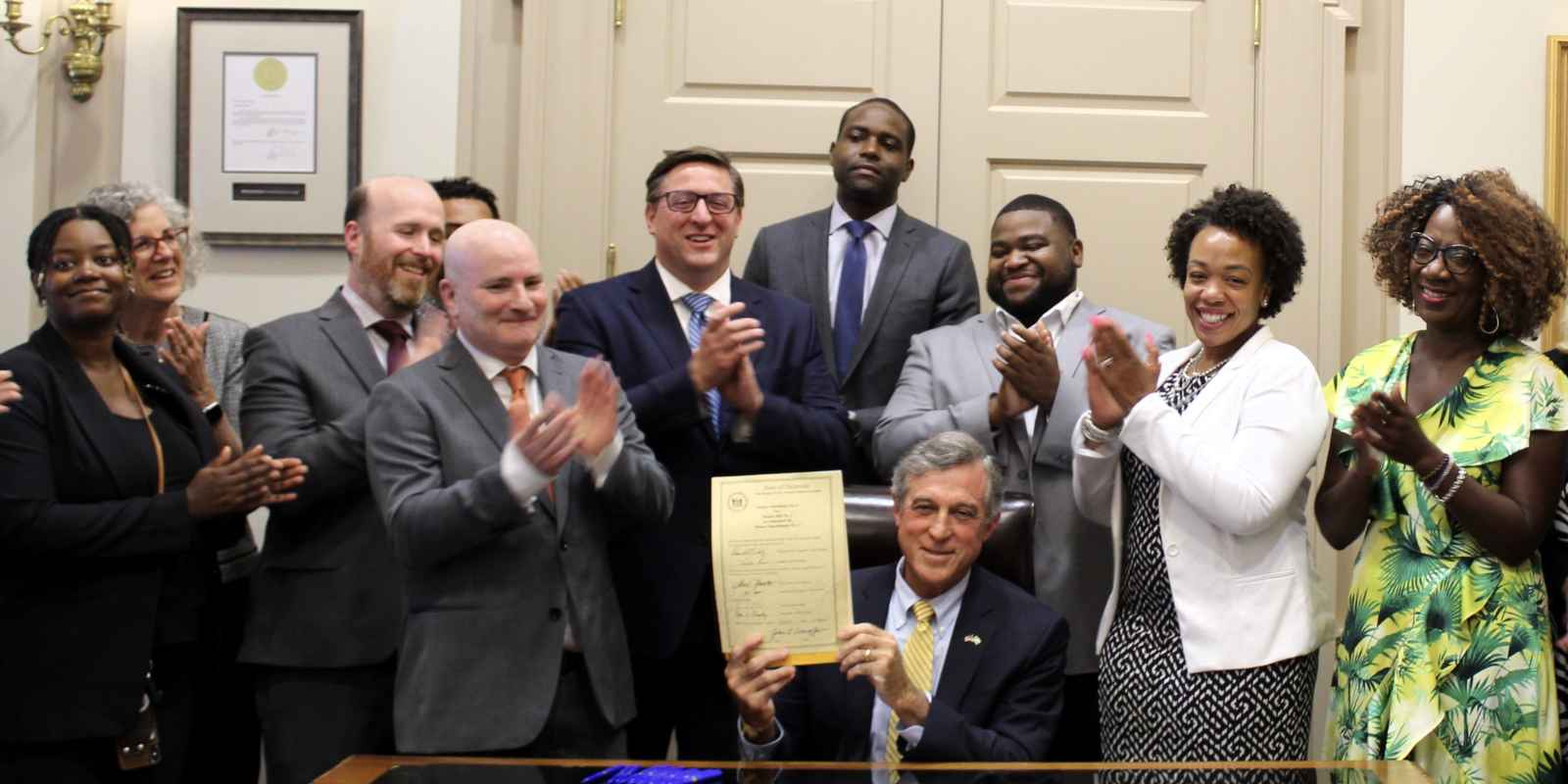 Gov. Carney sits at a desk holding the newly signed SS 1 for SB 1 legislation, surrounded by coalition partners and advocates.