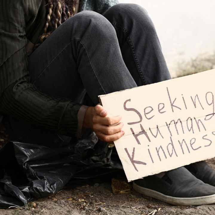 Person sitting on ground with cardboard sign that says "Seeking Human Kindness."