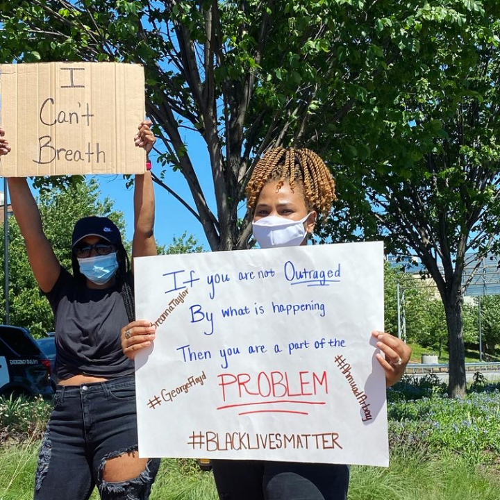 Two women holding signs at police violence protest, standing in front of Wilmington Police Department SUV on Martin Luther King Blvd in Wilmington